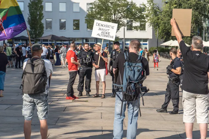 Demonstranten auf einem Platz halten ein Schild mit der Aufschrift „DEMOKRATIE BRAUCHT KEINE ALTERNATIVE“ hoch; eine Regenbogenfahne ist am Rand sichtbar.