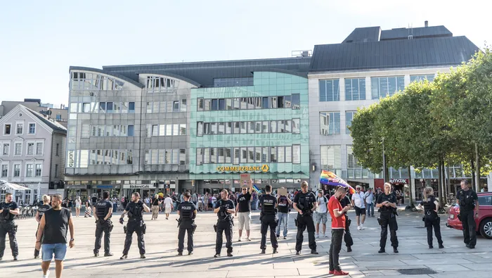 Mehrere Polizeibeamte in dunkler Uniform stehen auf einem öffentlichen Platz vor Geschäftsgebäuden.