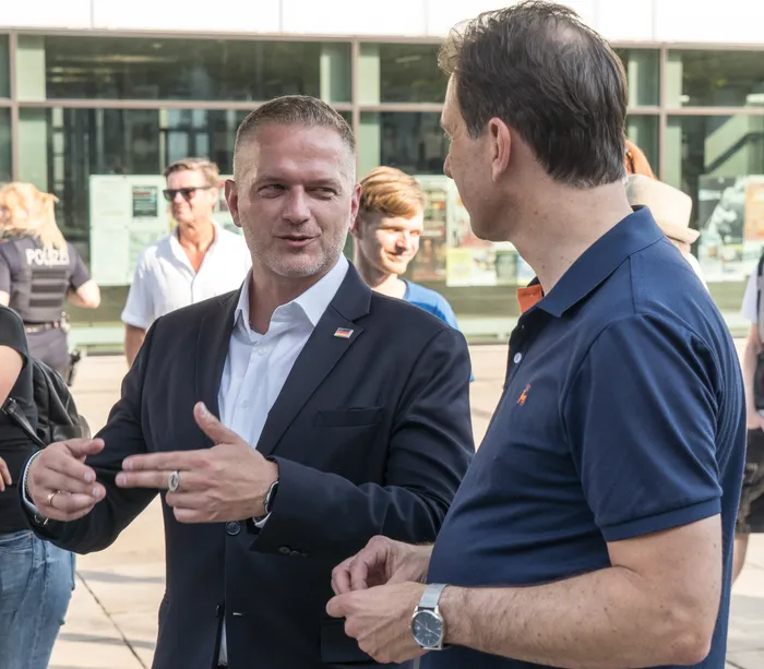 Adam Balten im dunklen Anzug gestikuliert während eines Gesprächs mit Christian Loose in einem blauen Poloshirt.