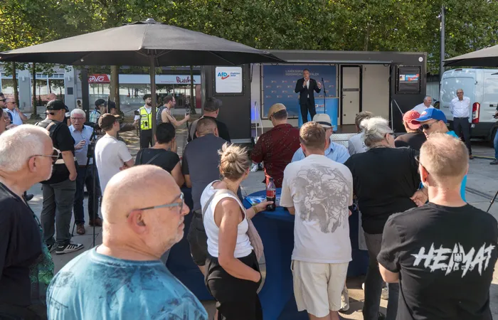 Ein Mann spricht an einem Rednerpult vor einem AfD-Banner vor einer Gruppe von Menschen im Freien.