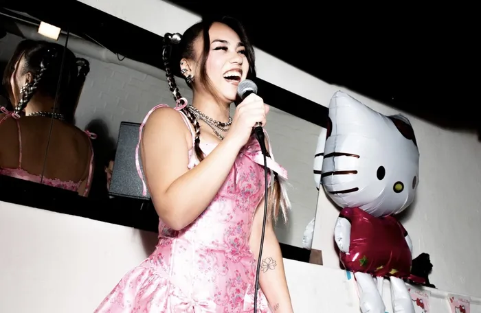 Sängerin Lean mit langen, geflochtenen Haaren singt in ein Mikrofon, während sie auf einer Bühne steht. Im Hintergrund ist eine große Hello-Kitty-Aufblasfigur zu sehen.