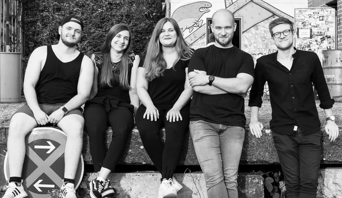 Fünf junge Erwachsene sitzen nebeneinander auf einer Mauer. Die drei Frauen und zwei Männer lächeln in die Kamera. Im Hintergrund ist eine bemalte Wand zu sehen.