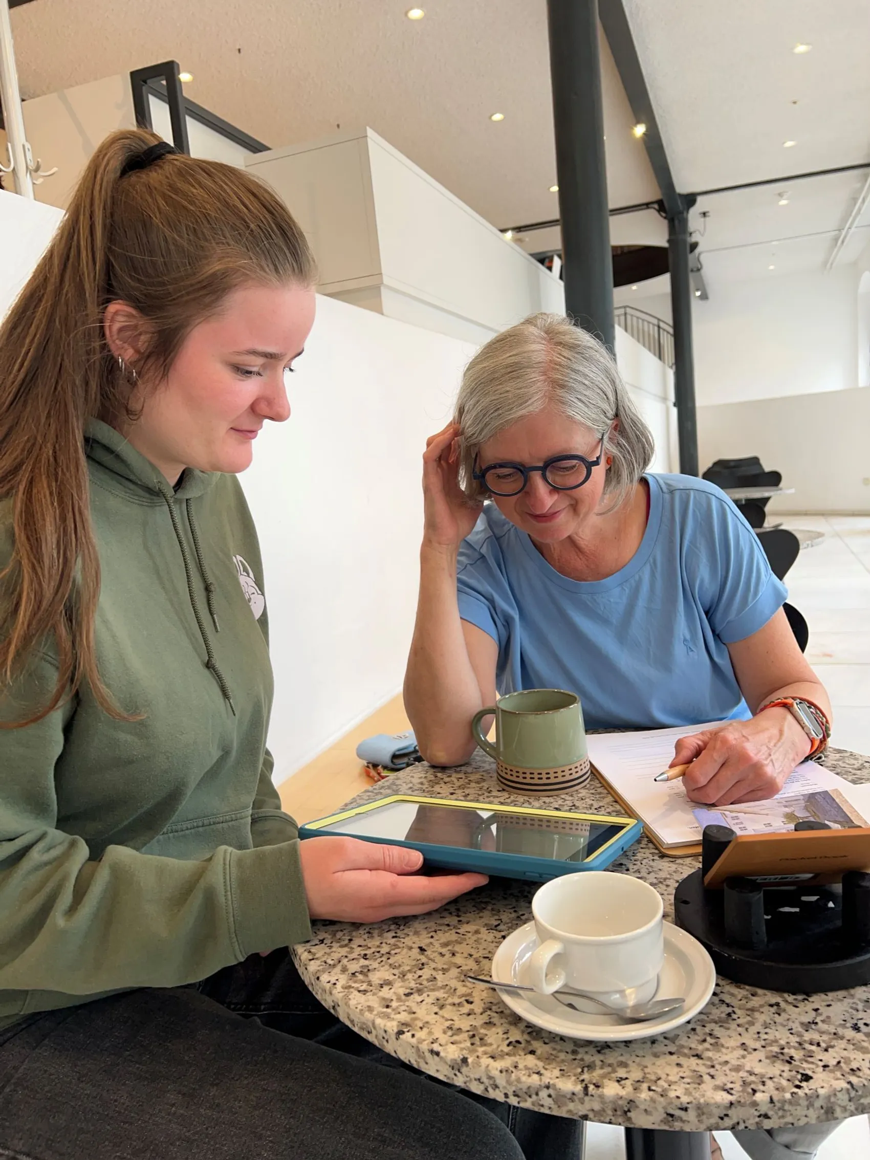 Eine junge Frau hält ein Tablet, während eine ältere Frau mit Brille in ein Notizbuch schreibt. Sie sitzen an einem Tisch mit Kaffeetassen.
