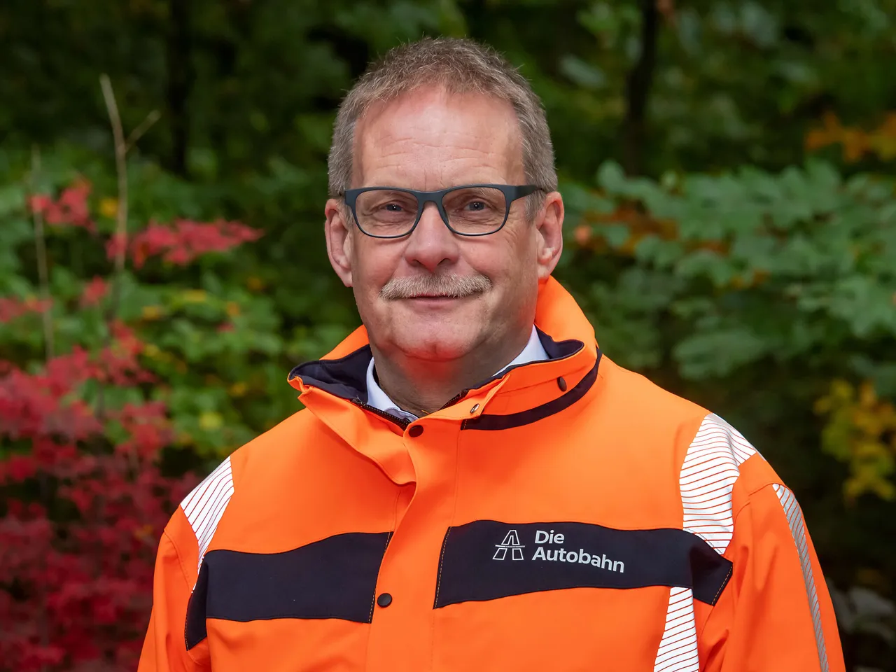 Porträt von Dirk Stiepert mit Brille und orangefarbener Arbeitsjacke mit dem Logo der Autobahn GmbH.