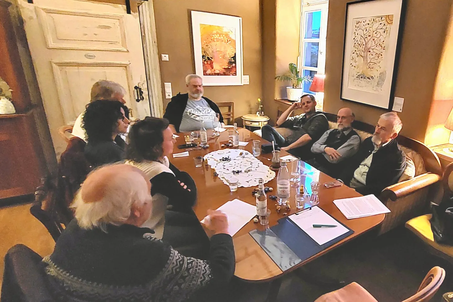 Eine Gruppe von sieben Personen sitzt um einen großen Holztisch in einem Wohnzimmer und diskutiert. Auf dem Tisch stehen Getränke und liegen Papiere bereit.