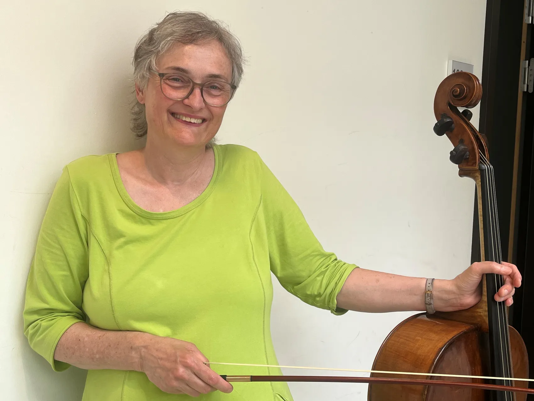 Eine Frau mit grauen Haaren und Brille lächelt in die Kamera, während sie ein Cello hält und den Bogen führt.