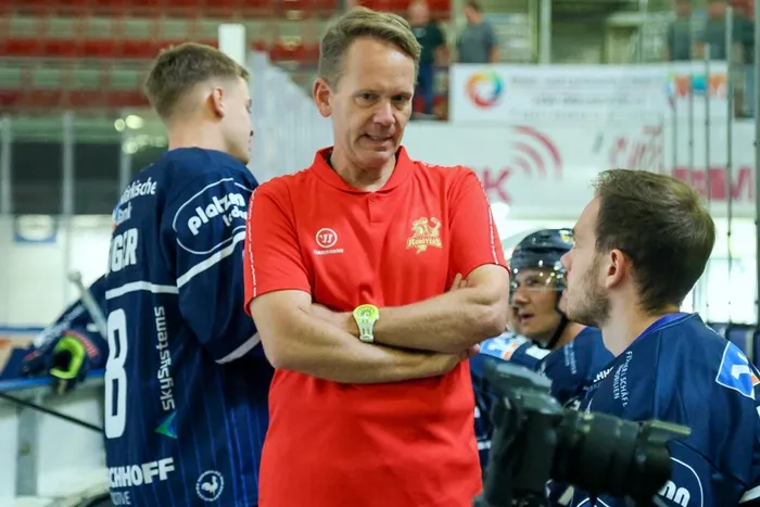 Dr. Jochen Veit steht mit verschränkten Armen in einem roten Poloshirt inmitten von Eishockeyspielern in blauen Trikots.