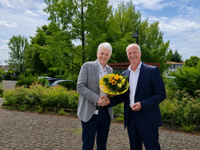 Zwei Männer im Anzug stehen lächelnd vor Bäumen und Büschen. Der linke Mann übergibt dem rechten einen Blumenstrauß mit gelben Sonnenblumen.