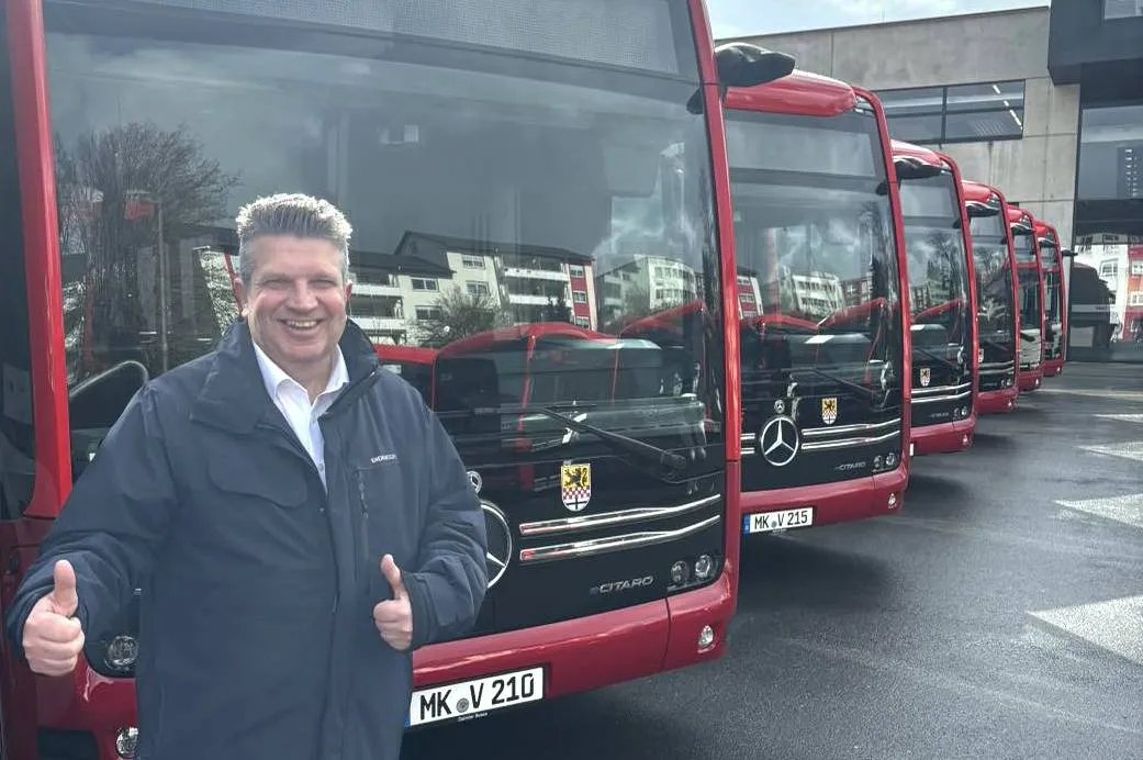 Gordan Dudas, Kandidat der SPD, steht lächelnd vor einer Reihe neuer roter Elektrobusse der MVG und zeigt den Daumen nach oben.