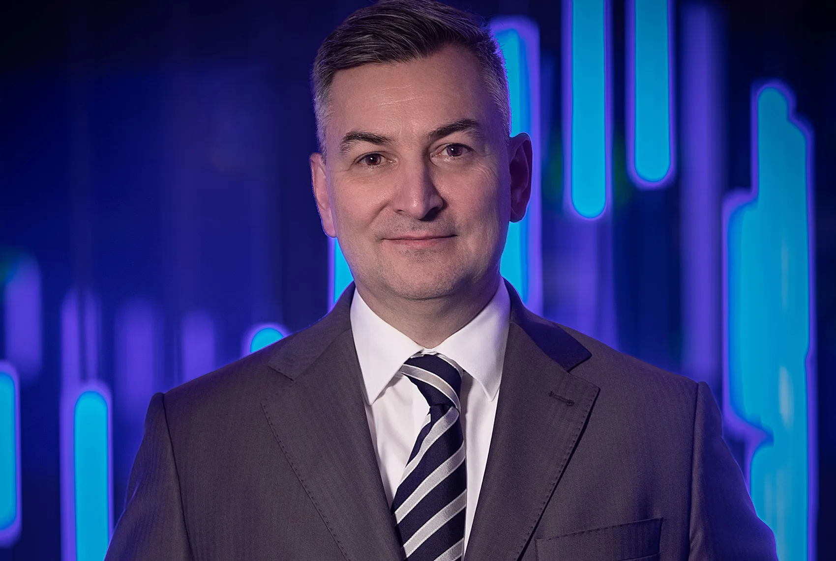 Markus Gürne in einem grauen Anzug und einer gestreiften Krawatte vor einem blau leuchtenden Hintergrund mit vertikalen Lichtakzenten.