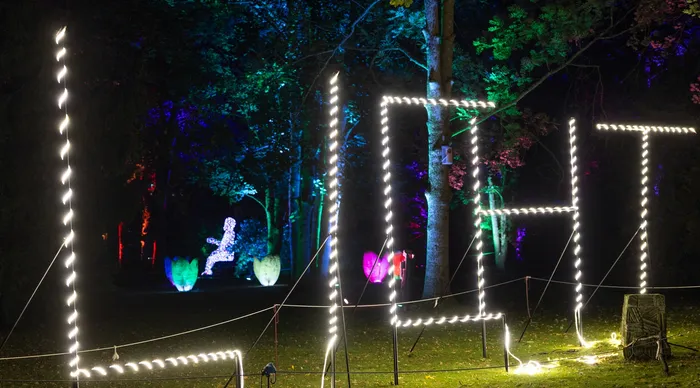 Große, mit Lichterketten gestaltete Buchstaben des Wortes „LIGHT“ vor farbig beleuchteten Bäumen und Lichtobjekten in einer nächtlichen Parklandschaft.