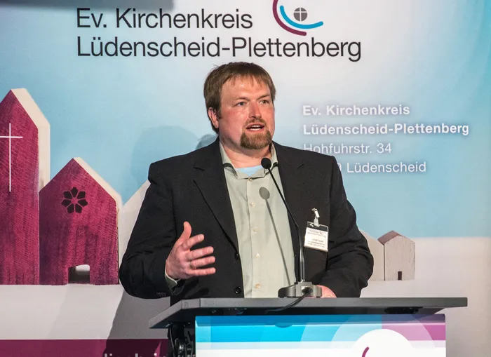 Johannes Seidel, Leiter des Kinder- und Jugendreferates, spricht auf der Kreissynode des Evangelischen Kirchenkreises Lüdenscheid-Plettenberg über die geplante neue Kinder- und Jugendvertretung. Er steht am Rednerpult vor einem Banner mit dem Logo und der Adresse des Kirchenkreises.