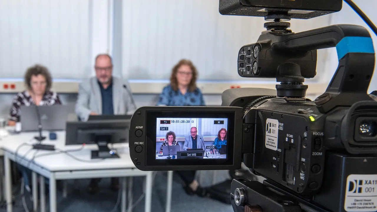 Eine Videokamera filmt eine digitale Sondersynode, bei der drei Personen an einem Tisch mit Laptops sitzen.