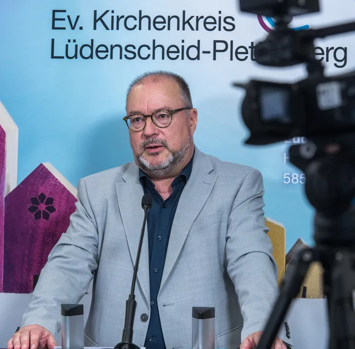 Superintendent Dr. Christof Grote spricht an einem Mikrofon vor einem blauen Hintergrund des Evangelischen Kirchenkreises Lüdenscheid-Plettenberg.