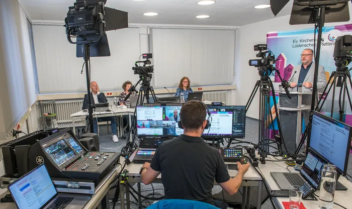 Ein Techniker bedient an einem Mischpult ein Studio-Setup mit Kameras und Monitoren für die Sondersynode vor einer Gruppe von Teilnehmern.