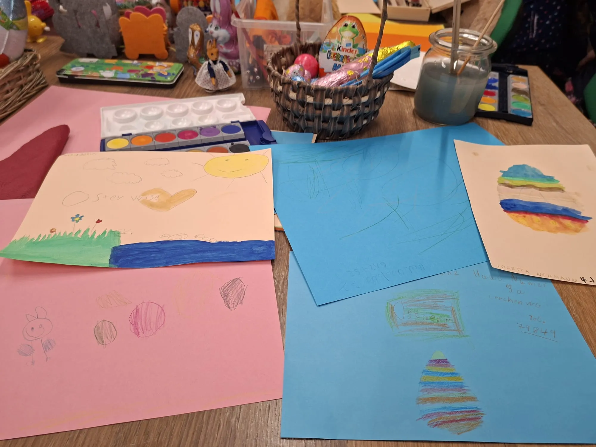 Kinderzeichnungen in verschiedenen Farben und Motiven liegen auf einem Tisch, darunter ein Osterhase und eine abstrakte Landschaft.
