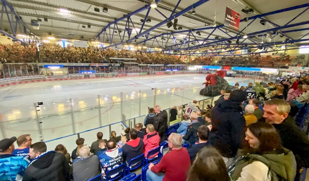 Innenansicht einer Eishalle mit Zuschauern auf den blauen Sitzen und einer Eisfläche, auf der sich zwei Eishockeyspieler befinden.