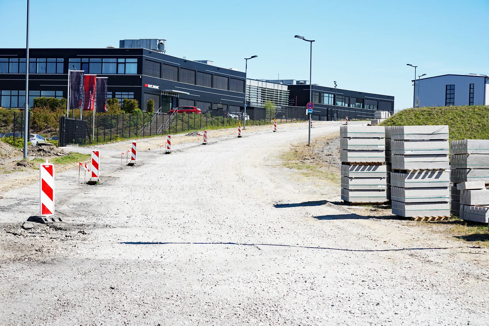 Eine Baustelle vor einem modernen Gewerbegebäude mit dem Schriftzug „ELECTRONIC“. Sicherheitskegel und Betonblöcke markieren den Weg.