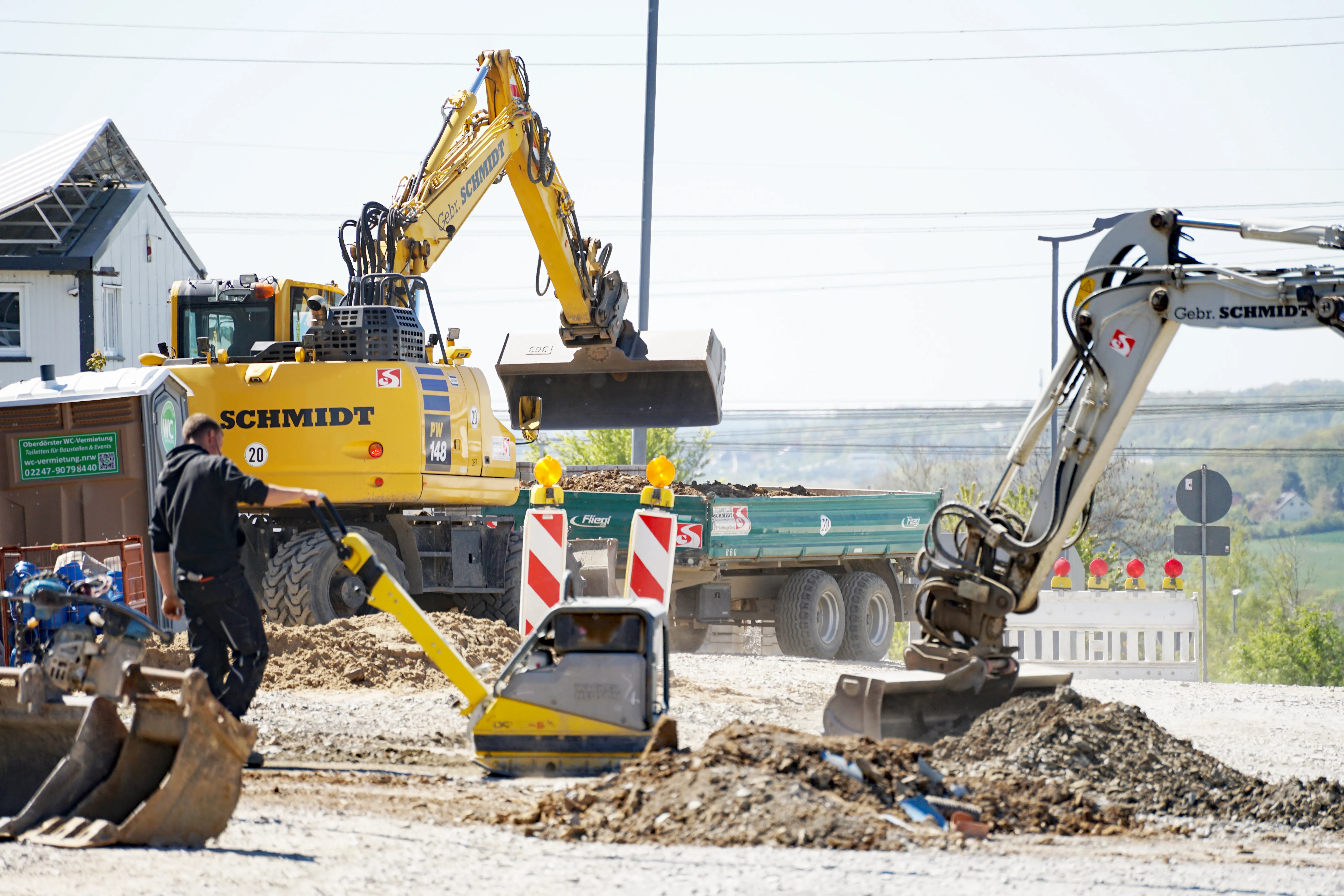 Zwei Bagger und ein LKW entladen Erdreich auf einer Baustelle. Ein Bauarbeiter ist mit Schaufel im Vordergrund zu sehen.