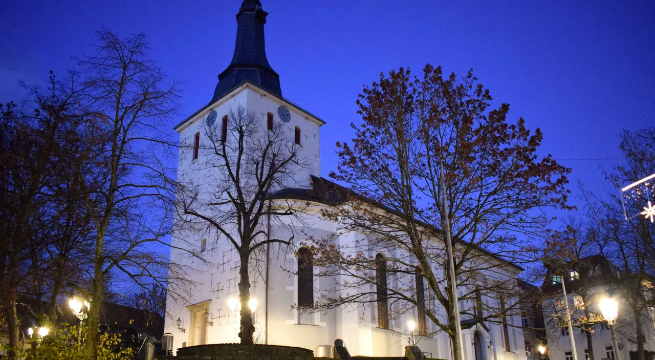 Die weiß beleuchtete Erlöserkirche mit dunklem Turm vor einem blauen Abendhimmel, umgeben von Bäumen.