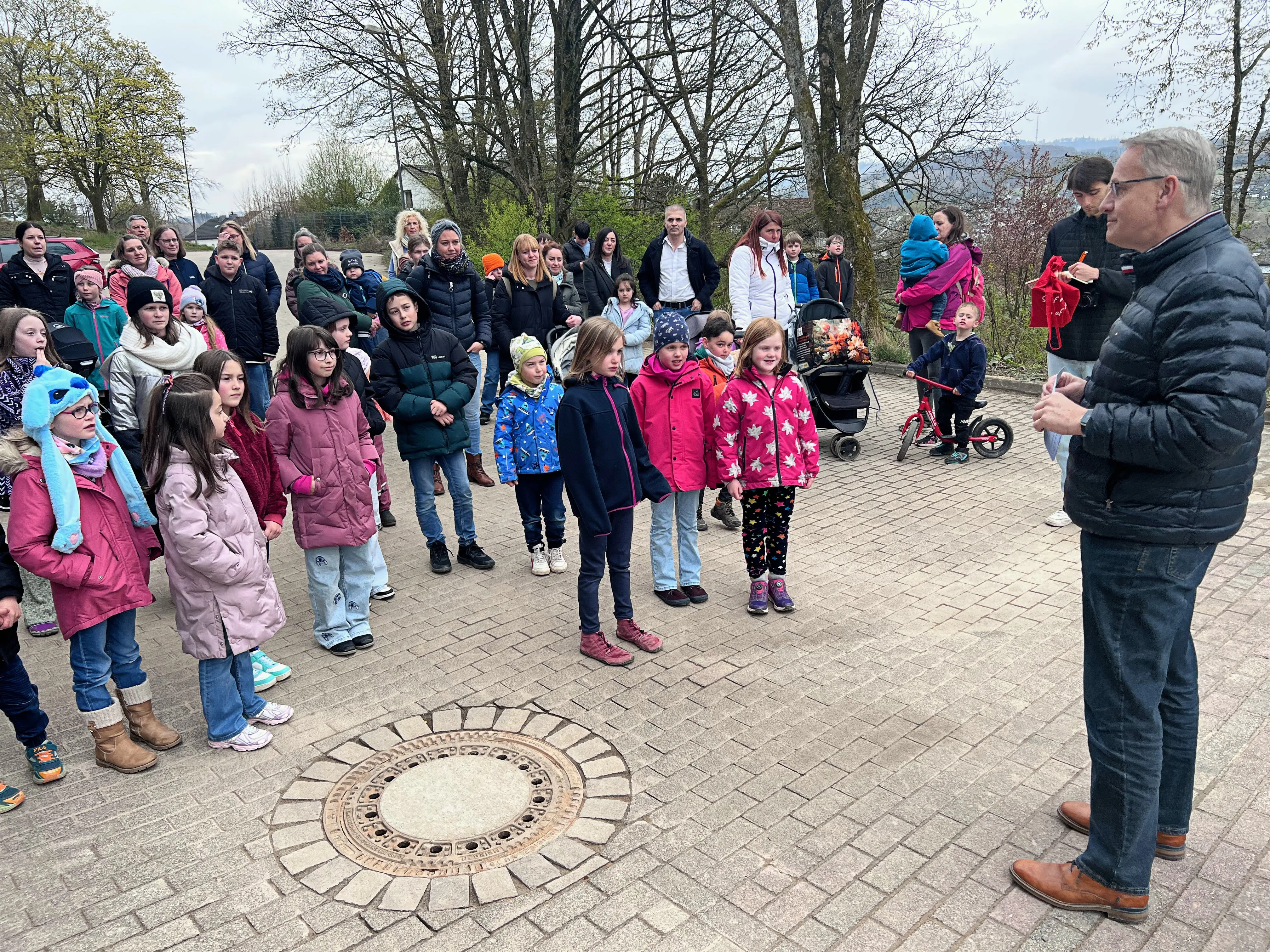 Eine Gruppe Kinder und Erwachsene steht im Kreis um ein dekoratives Element im Pflaster eines Spielplatzes, während ein Mann spricht.