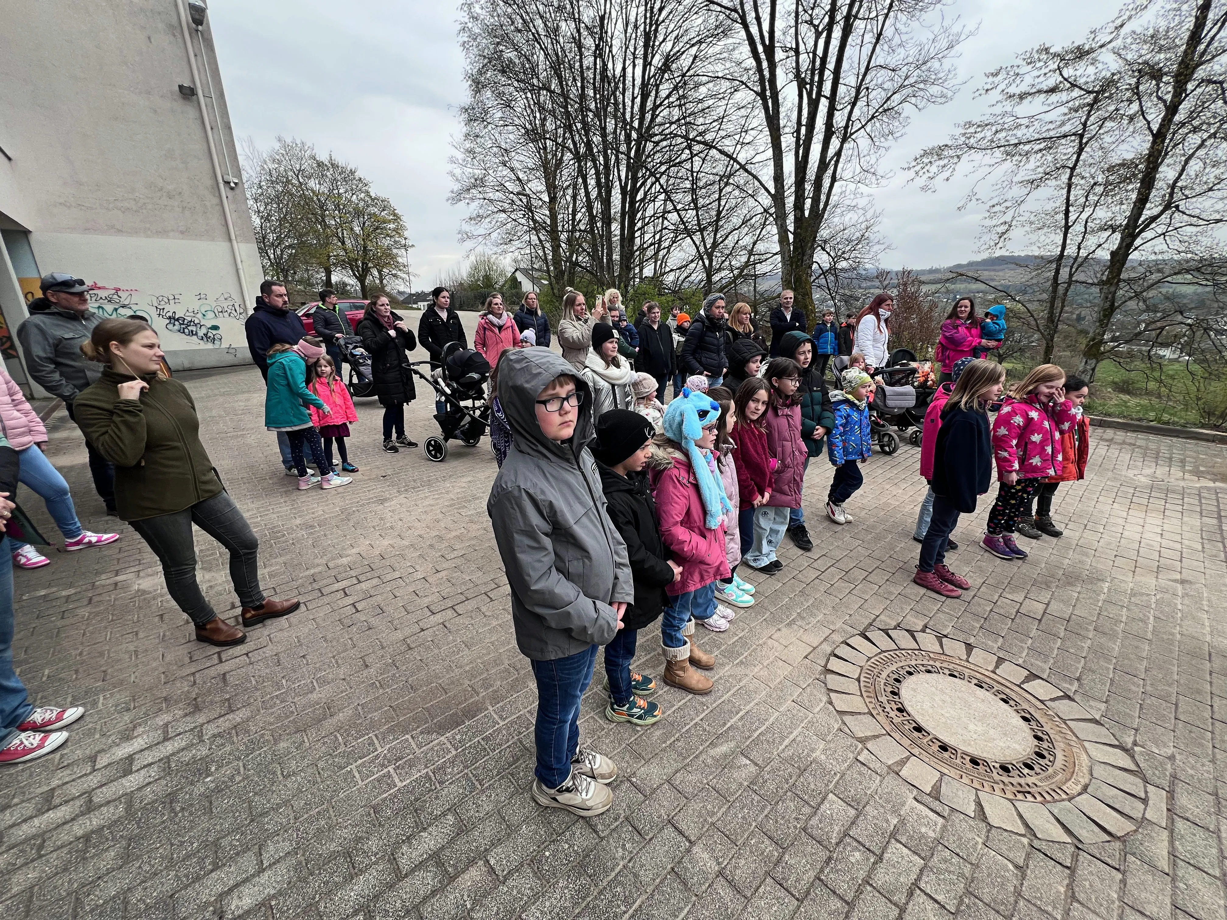 Eine Gruppe Kinder und Eltern steht auf einem gepflasterten Platz vor einem Spielplatz. Im Hintergrund sind Bäume und ein Gebäude zu sehen.