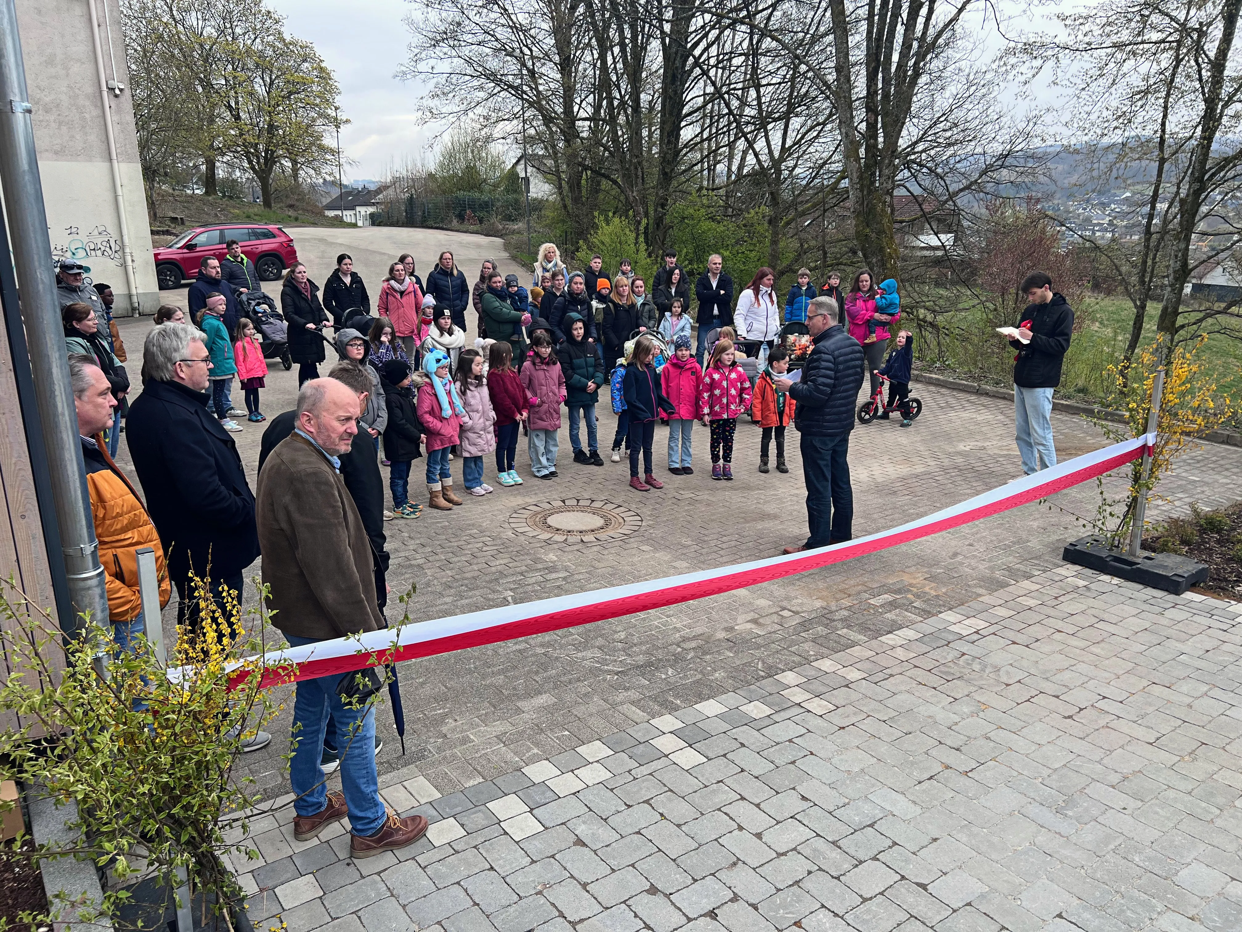 Eine Gruppe von Erwachsenen und Kindern steht um einen Baum, dessen Stamm mit einem rot-weißen Band umwickelt ist. Im Hintergrund sind weitere Personen und ein Wohngebiet zu sehen.