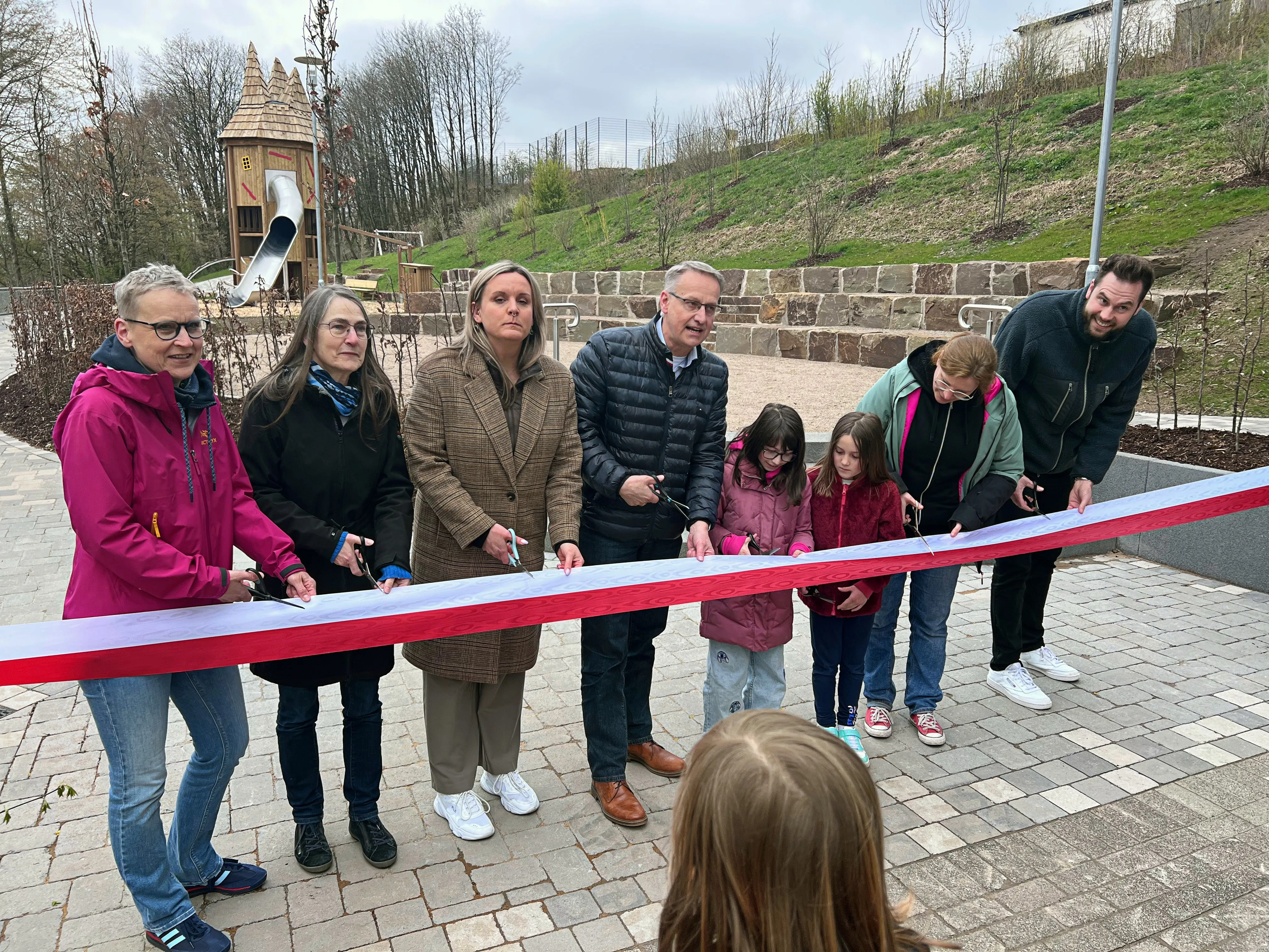 Eine Gruppe von Erwachsenen und Kindern schneidet ein rotes Band durch, um einen neuen Spielplatz zu eröffnen. Im Hintergrund ist ein Spielturm zu sehen.