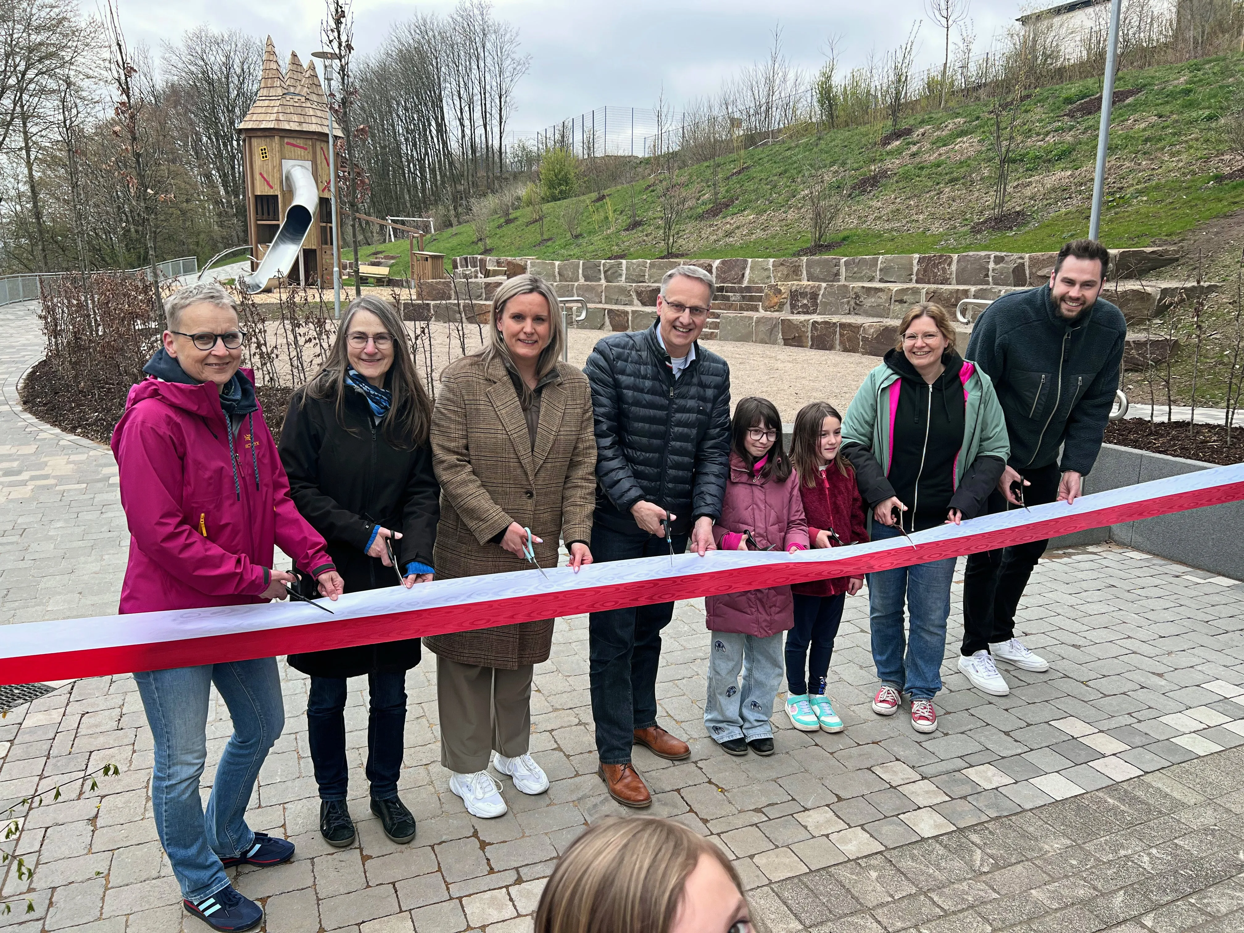 Gruppe schneidet rotes Band vor neuem Spielplatz mit Holzburg im Hintergrund durch.