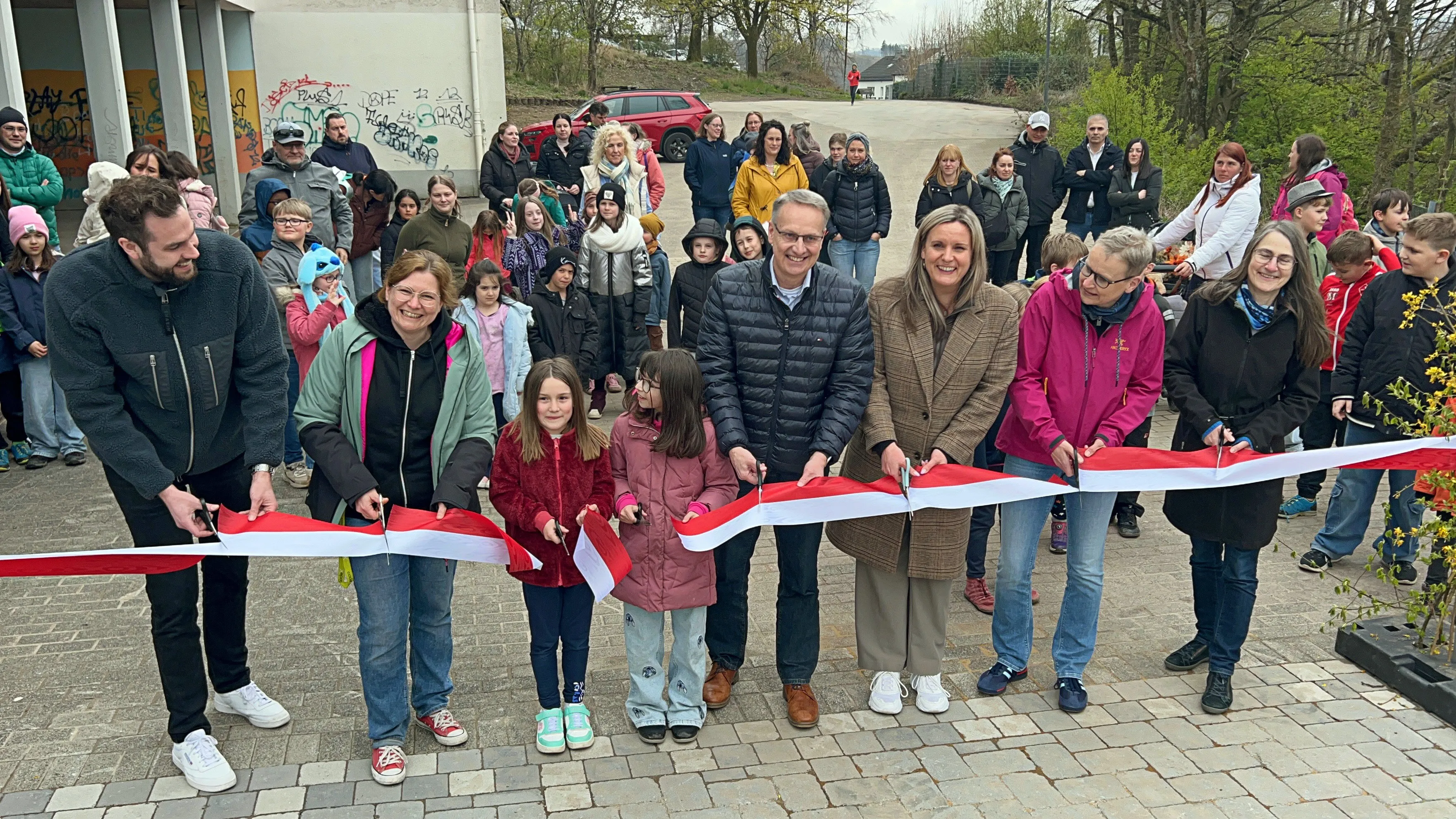 Eine Gruppe von Erwachsenen und Kindern schneidet ein rotes und weißes Eröffnungsband vor einem Spielplatz durch.