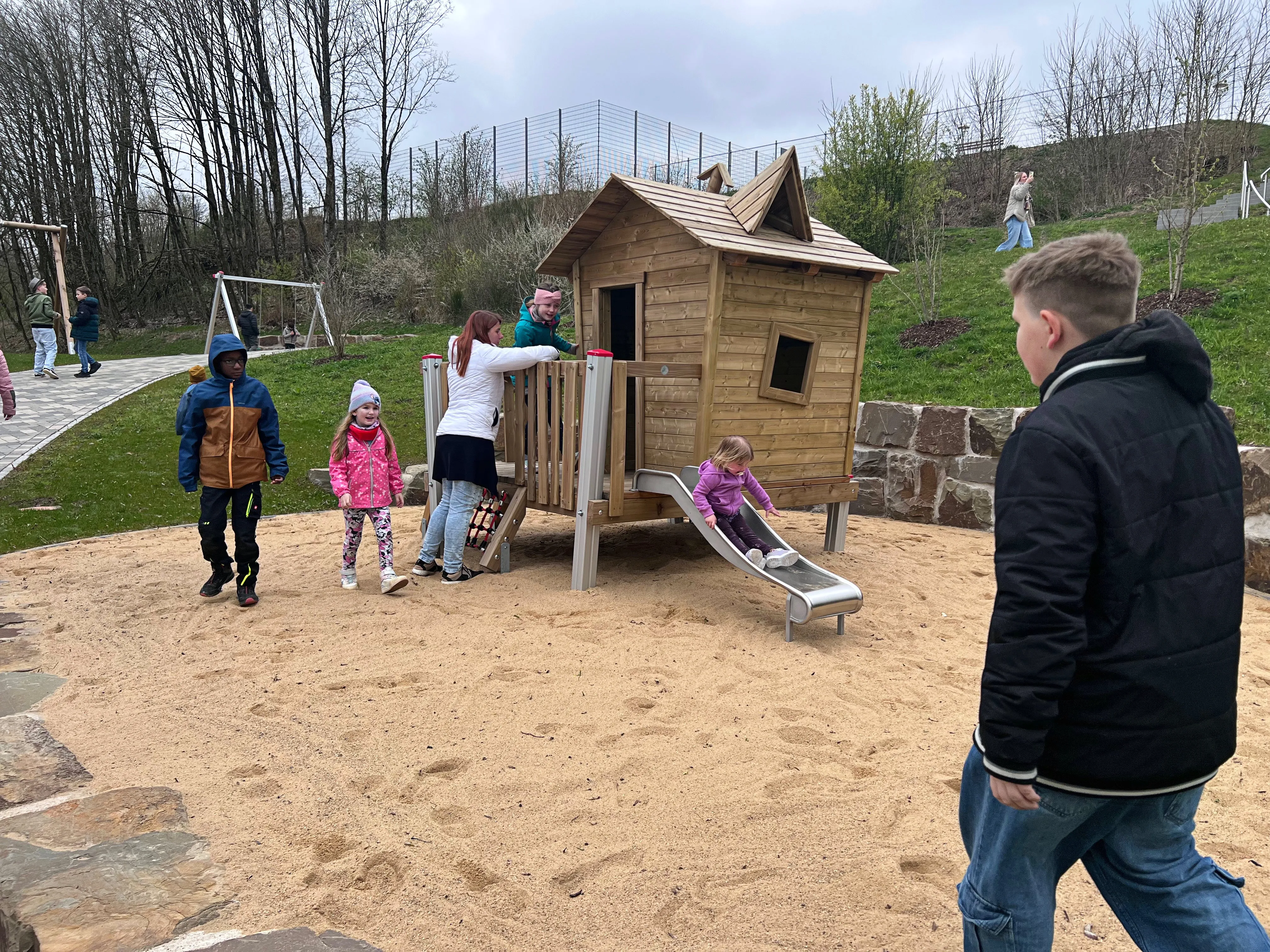 Kinder spielen auf einem neuen Spielplatz mit einem kleinen Holzhaus und einer Rutsche, während ein Junge im Vordergrund wegschaut.