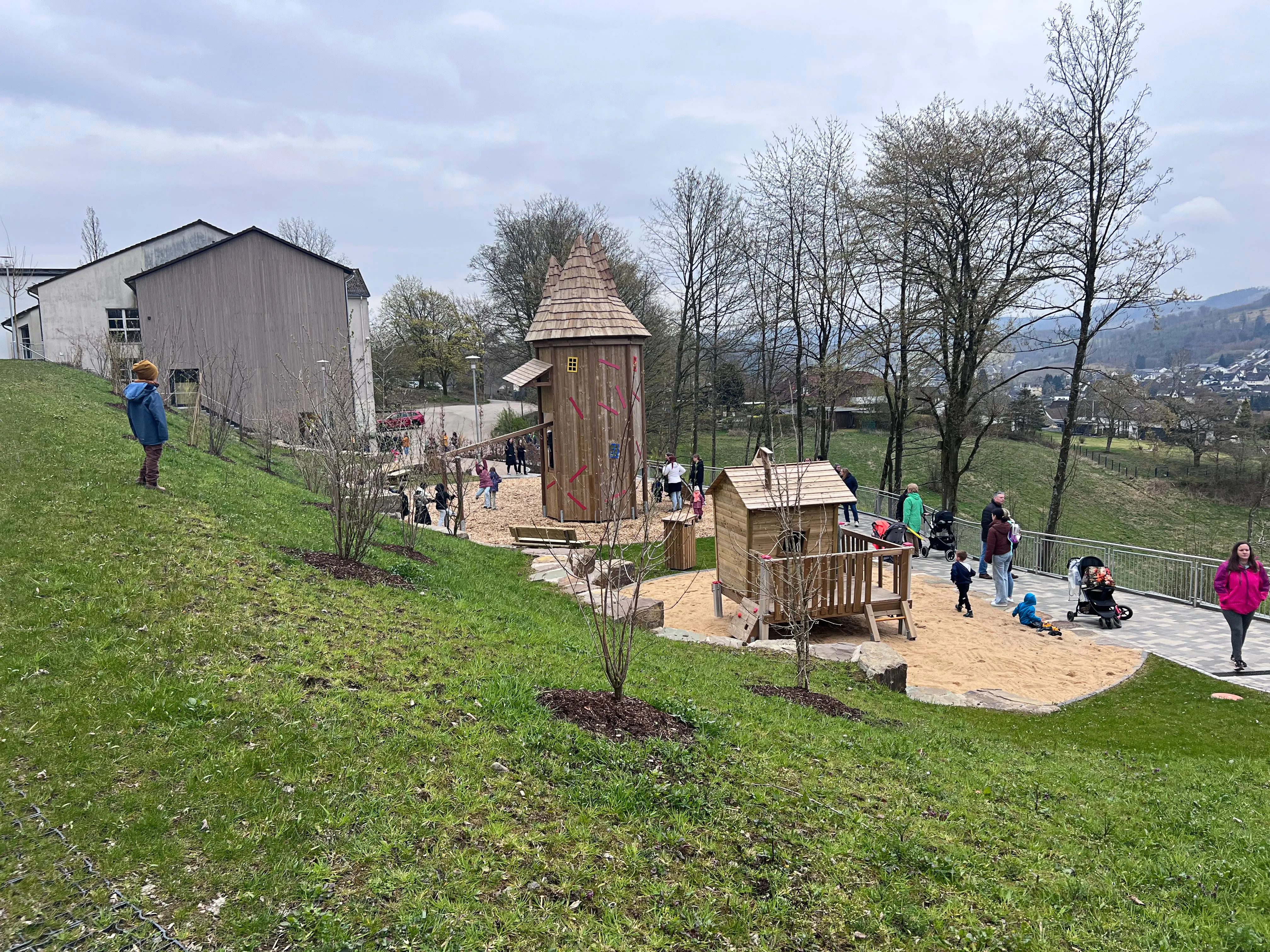 Ein Spielplatz mit hölzernen Spielstrukturen und Sandfläche, umgeben von einer Rasenfläche und Bäumen. Im Hintergrund ein Gebäude und eine Hügellandschaft.