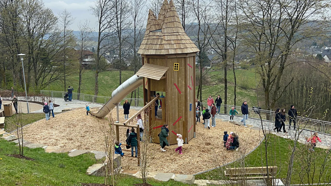 Kinder spielen auf einem neuen Spielplatz mit einem hölzernen Turm und Rutsche, umgeben von Rasen und Bäumen.