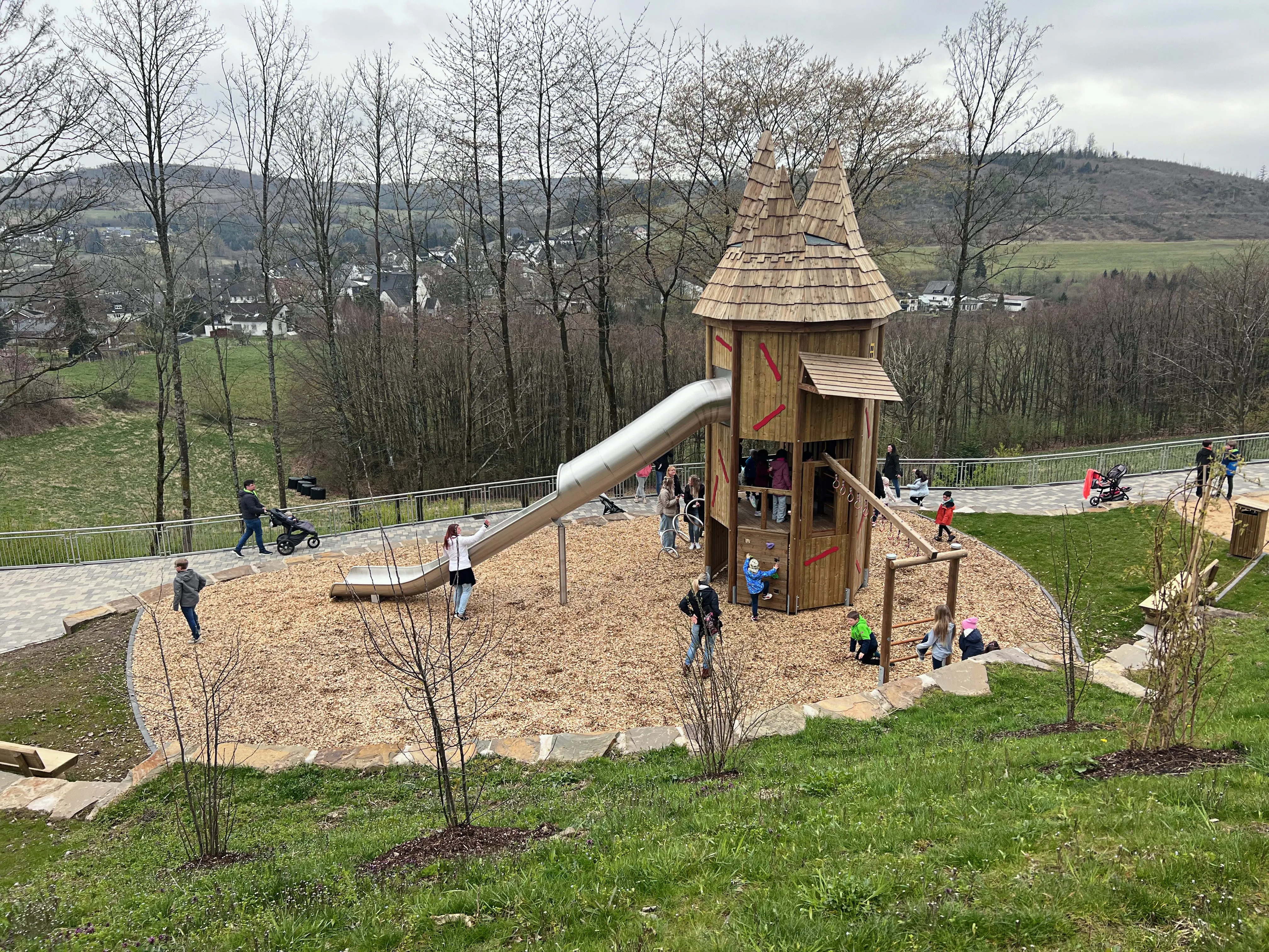 Ein Spielturm mit Rutsche steht auf einem mit Rindenmulch belegten Spielplatz. Kinder spielen und erkunden die Anlage, im Hintergrund eine Wohnsiedlung und bewaldete Hügel.