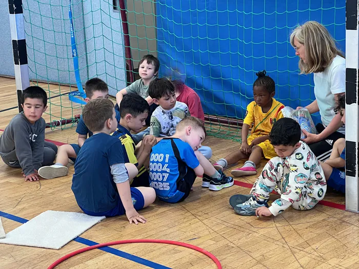 Eine Gruppe von Kindern sitzt im Kreis auf dem Hallenboden und lauscht einer Frau, die im Hintergrund steht. Die Kinder sind unterschiedlich gekleidet, einige tragen Sportkleidung.