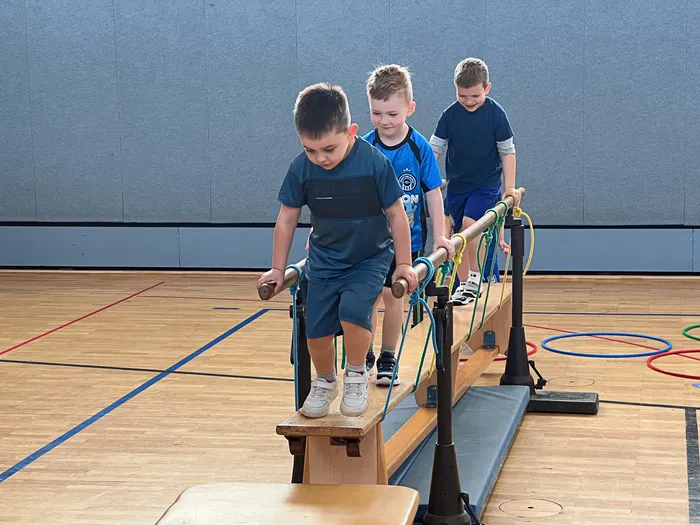 Drei Jungen balancieren auf einer schmalen Holzkonstruktion mit seitlichen Stangen in einer Turnhalle. Sie tragen Sportkleidung und konzentrieren sich auf ihre Aufgabe.