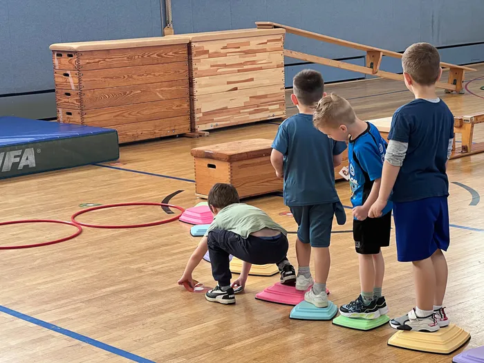 Vier Jungen stehen und hocken auf bunten, erhöhten Flächen in einer Turnhalle. Im Hintergrund sind weitere Sportgeräte und eine Holzkonstruktion zu sehen.