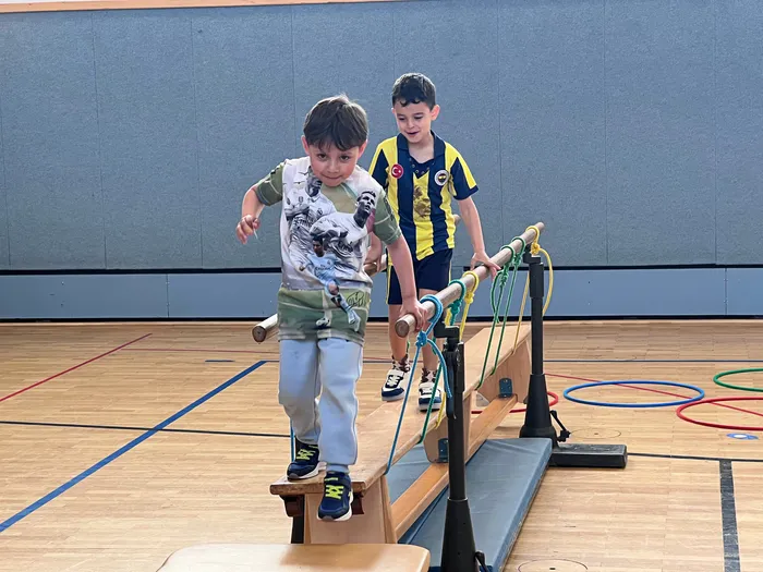 Zwei Jungen balancieren auf einem selbstgebauten Balken in einer Turnhalle. Der Junge im Vordergrund trägt ein T-Shirt mit Tiermotiven und helle Schuhe, während der andere einen Fußballtrikot trägt.
