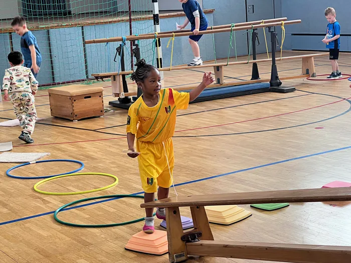 Ein junges Mädchen mit dunkler Hautfarbe und geflochtenem Haar steht in einer Turnhalle auf einer kleinen Holzkonstruktion und hebt die Hand. Im Hintergrund sind andere Kinder bei Übungen zu sehen.