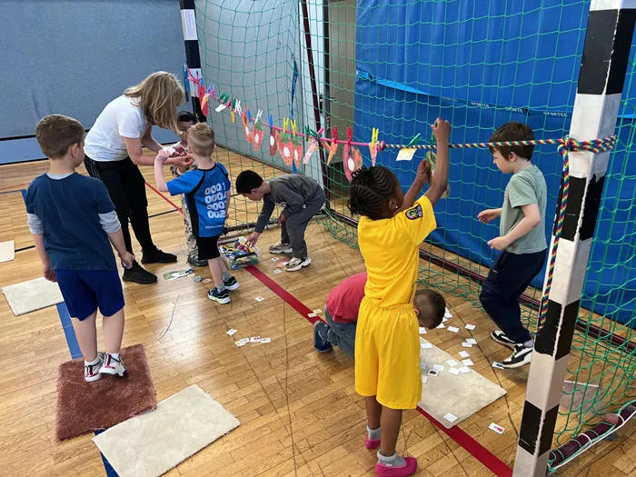 Kinder sammeln in einer Turnhalle Kärtchen vom Boden auf. Einige Kinder werden von einer Frau beaufsichtigt. Im Hintergrund eine blaue Wand mit bunten Bändern.