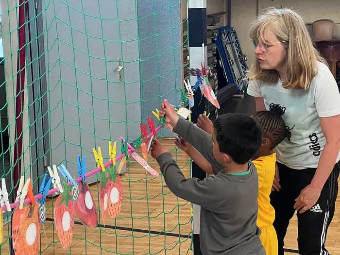 Eine Frau und ein Kind befestigen bunte Papierarbeiten mit Wäscheklammern an einer Schnur vor einem grünen Netz. Im Hintergrund sind weitere Kinderutensilien und ein Sportraum zu sehen.