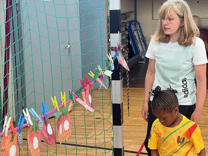 Eine Frau und ein Kind betrachten bunte, mit Wäscheklammern befestigte Papierarbeiten an einem Netz in einer Turnhalle. Im Hintergrund sind weitere Sportgeräte und eine Tür zu sehen.