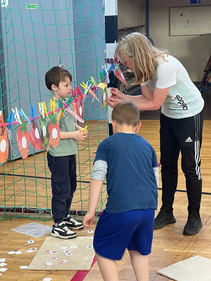 Eine Frau hilft drei Kindern bei einem Spiel auf dem Boden in einer Turnhalle. Im Hintergrund sind bunte Bastelarbeiten an einem Netz zu sehen.