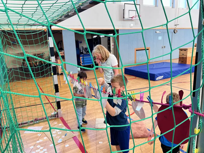 Kinder befestigen Wäscheklammern an einem Netz in einer Turnhalle, während eine Frau im Hintergrund zusieht. Im Hintergrund sind Sportgeräte und ein Basketballkorb zu erkennen.