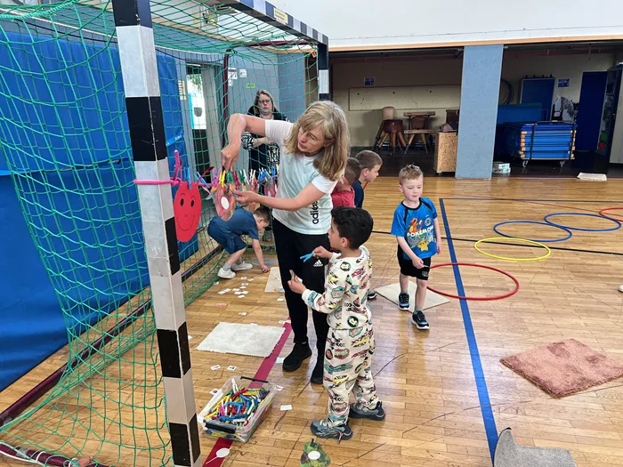 Eine Frau mit grauen Haaren hilft einem Jungen in Schlafanzug beim Spielen mit bunten Bändern in einer Turnhalle. Im Hintergrund sind weitere Kinder und eine Frau zu sehen.