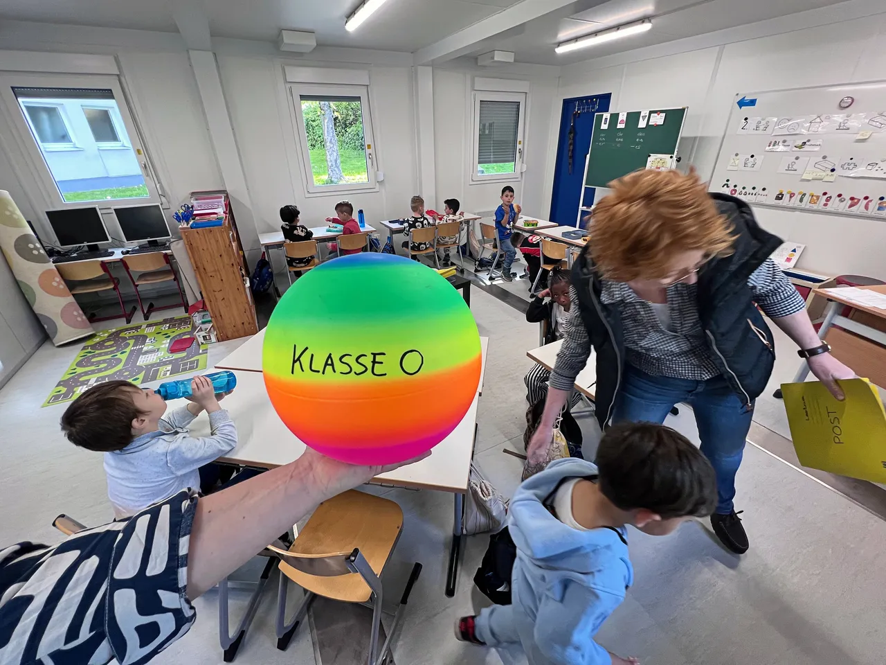Kinder sitzen und stehen um einen großen, bunten Ball mit der Aufschrift „KLASSE 0“ in einem hellen Klassenzimmer. Eine Lehrerin steht daneben und deutet auf den Ball.
