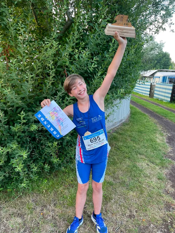 Mats Emil Wolff in blauer Sportkleidung hält lächelnd eine Urkunde und einen Holzpokal in die Höhe.