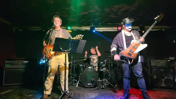 Eine Band tritt bei dem Benefizkonzert „Rock for Peace“ auf einer Bühne auf. Zu sehen sind ein singender Gitarrist, ein Schlagzeuger und ein maskierter Bassist.