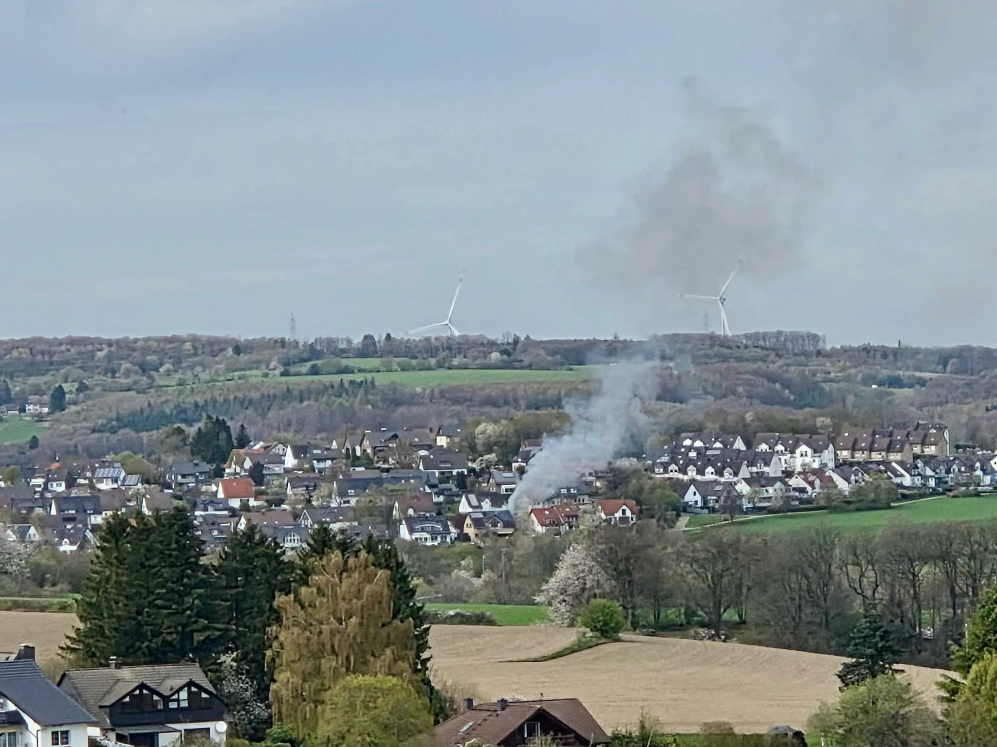 Rauch steigt über einem Wohngebiet auf, im Hintergrund Windräder und bewaldete Hügel.