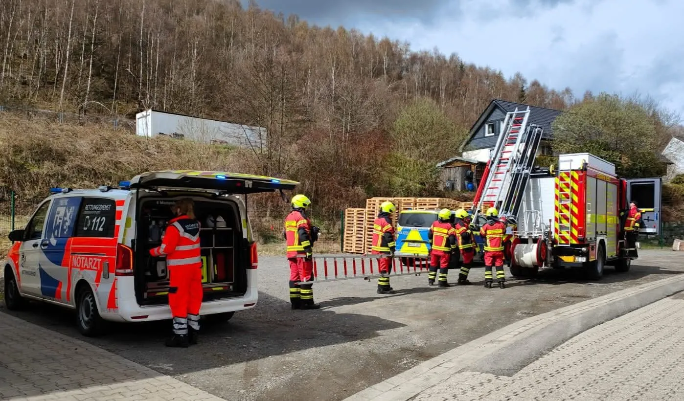 Feuerwehrleute stehen vor einem geöffneten Notarzteinsatzwagen und einem Einsatzwagen mit Leiter, während sie an einem Wohnhaus arbeiten.